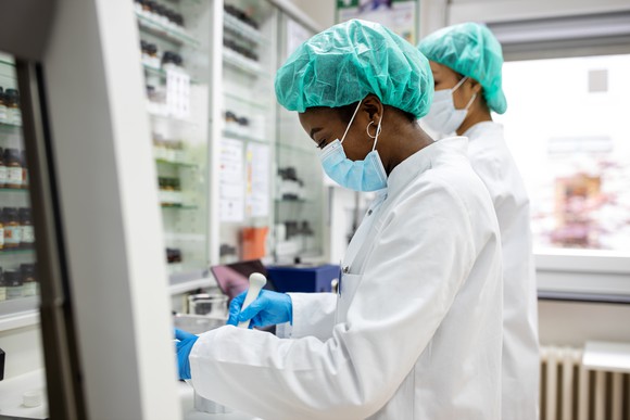 Black female laboratory worker, wearing a mask, hairnet, white coat, and gloves, handles laboratory equipment beside a coworker.
