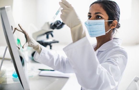 Woman with dark hair and skin. wearing a surgical mask and a white lab coat holds up a vial in one hand, with her other hand working on a touch-screen device in a laboratory setting. 