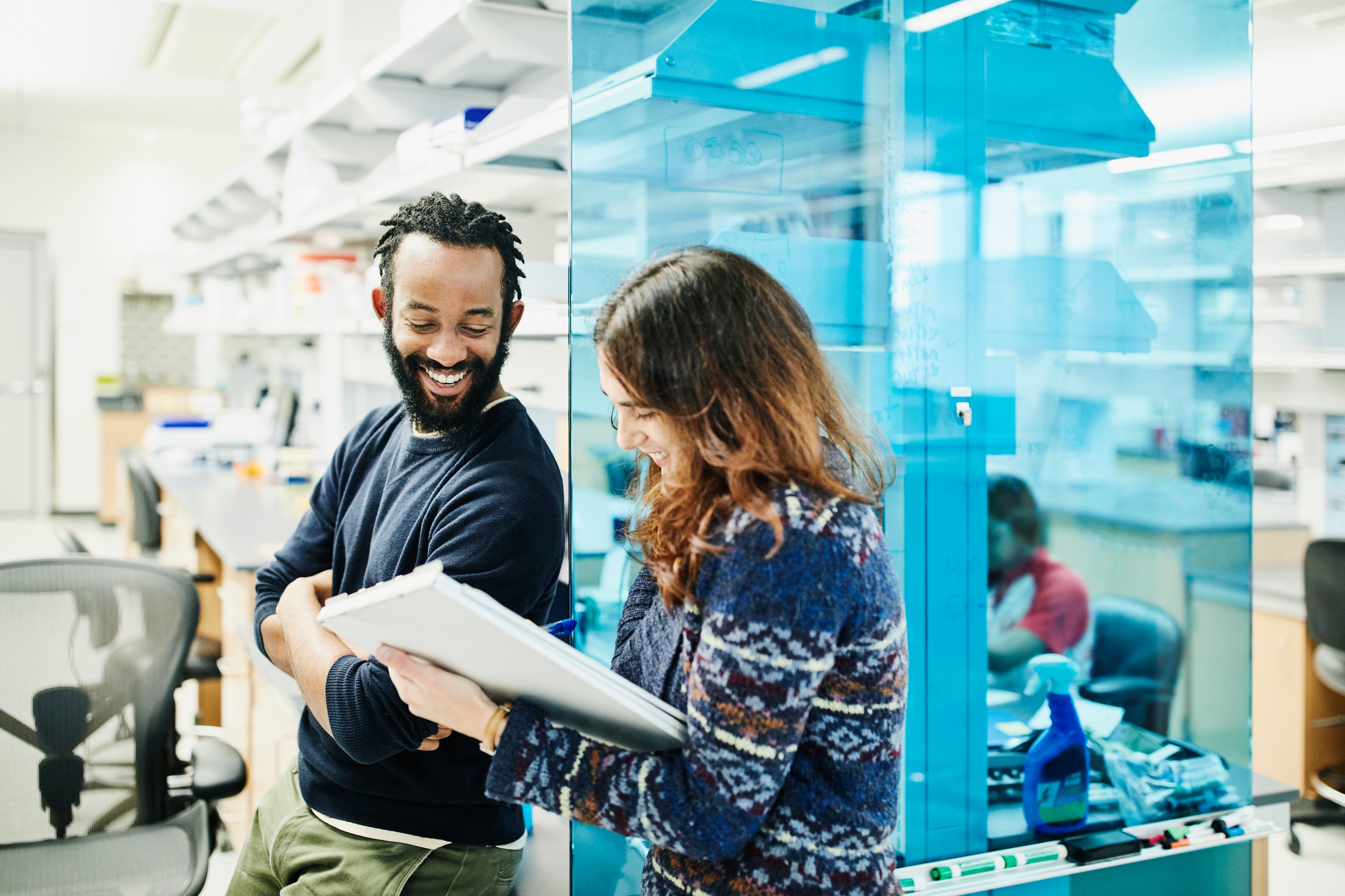 Man and woman, both wearing casual clothing, smile while looking at a pad of paper while working in a laboratory setting.