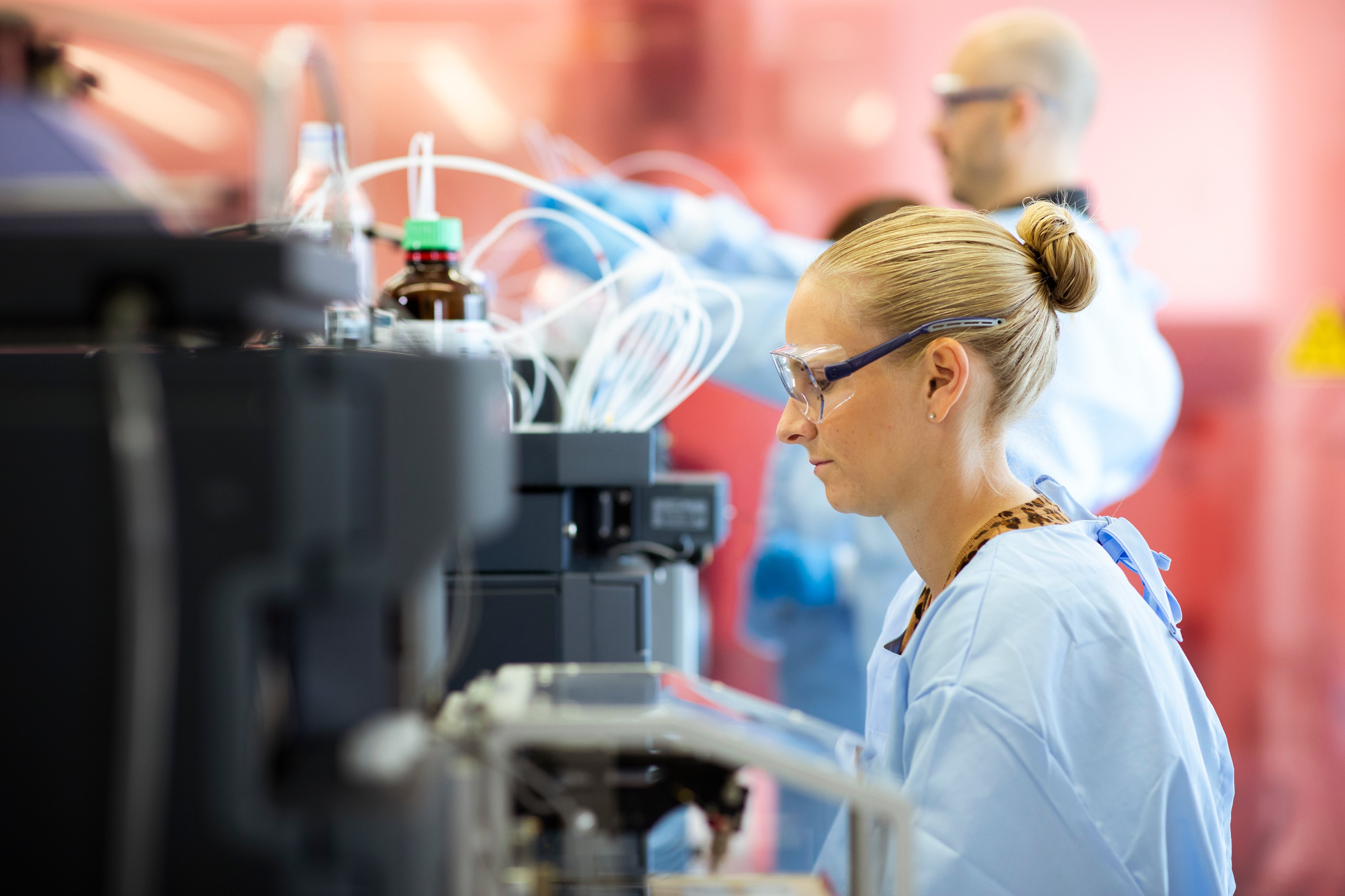 Blonde, white woman with hair pulled back in a bun, wearing personal protective gown and goggles, works with a large grey machine in a laboratory setting.