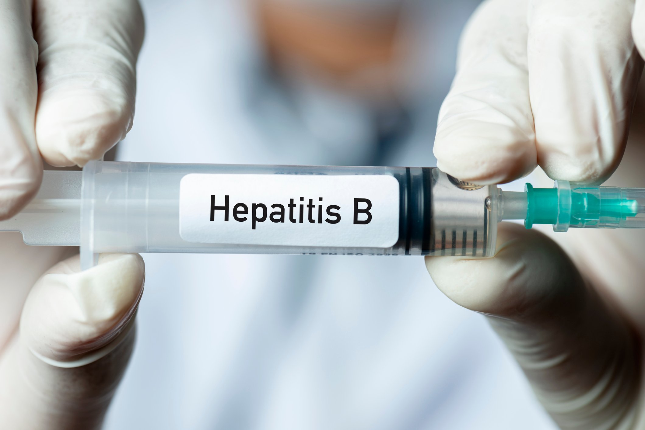 Gloved fingers holding a syringe with a label stating "Hepatitis B."