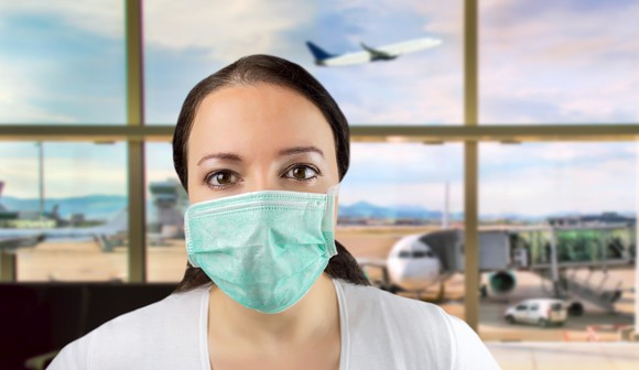A woman wearing a surgical mask at an airport, with a plane taking off behind her