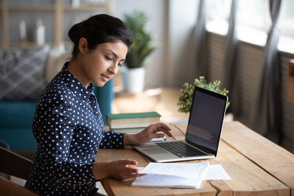 A woman glances as notes as she sits in front of her laptop.