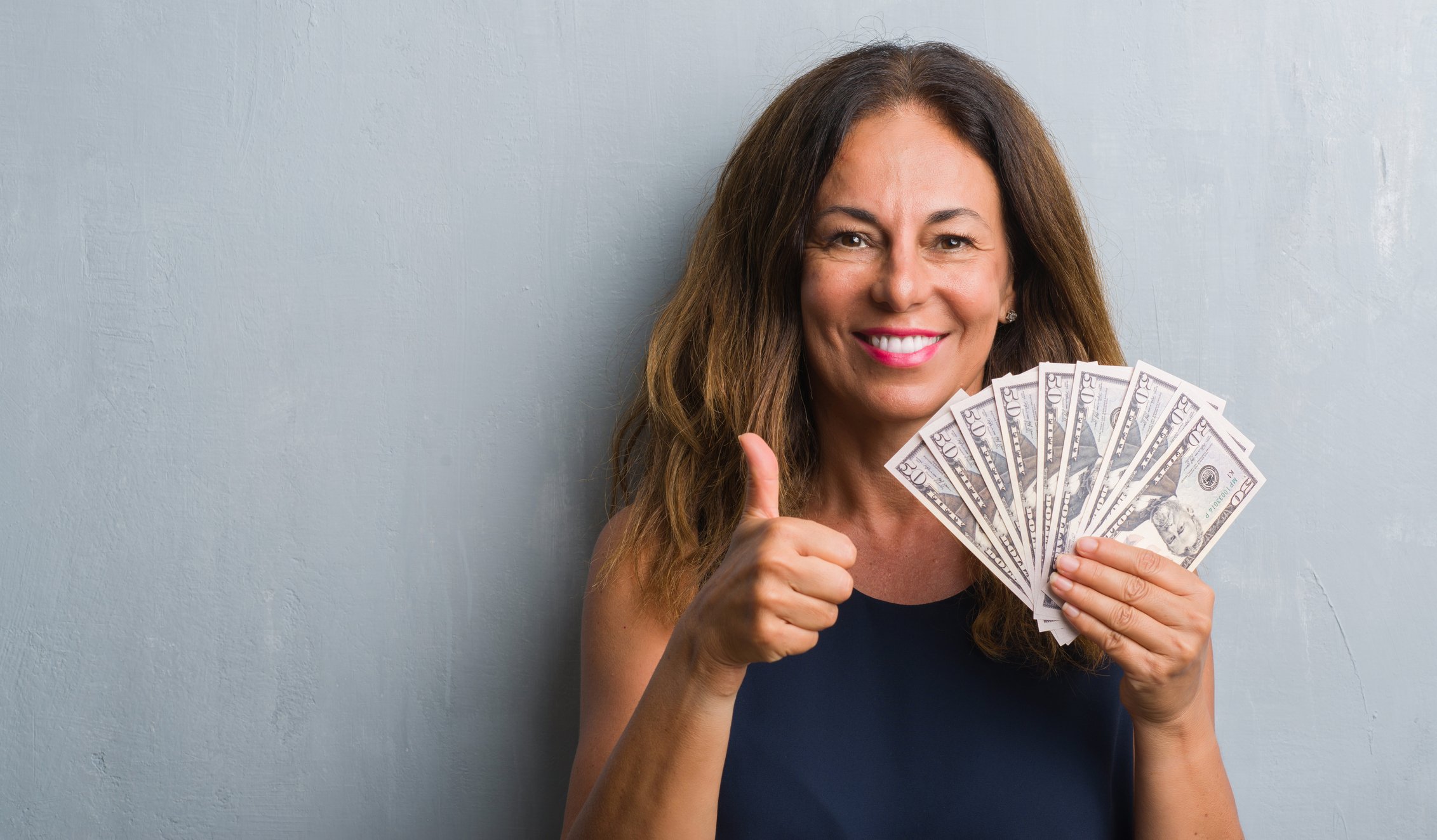 A smiling woman holding hundred dollar bills fanned out is giving a thumbs-up.