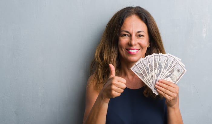 A smiling woman holding hundred dollar bills fanned out is giving a thumbs-up.
