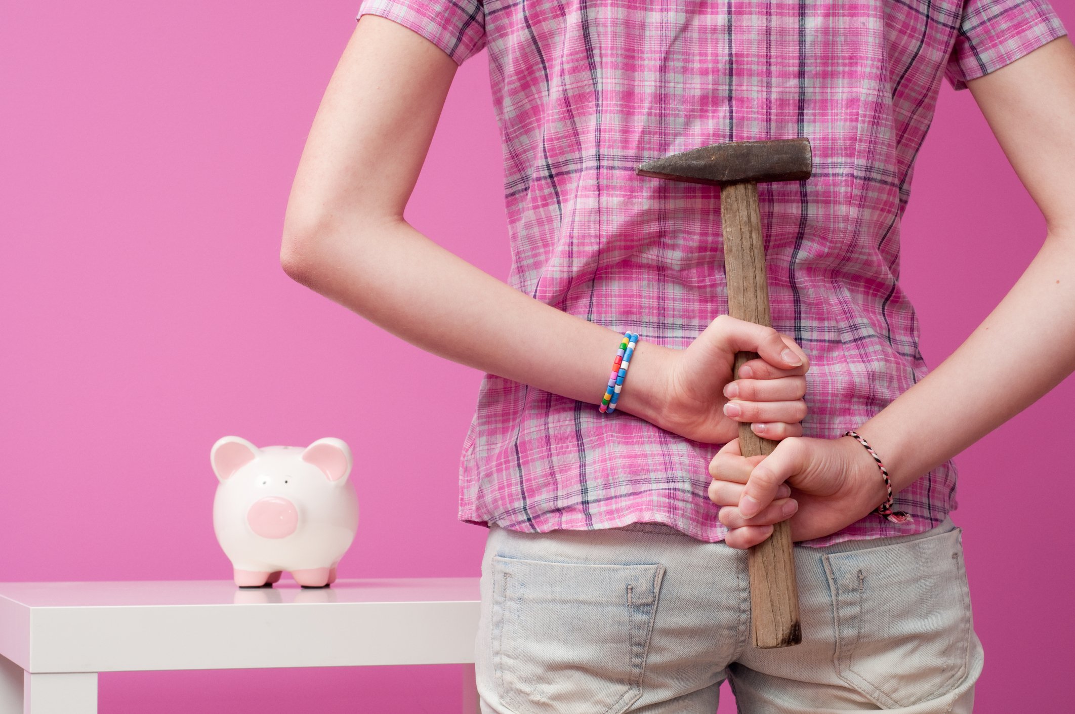 A person holds a hammer behind their back while looking toward a piggy bank.