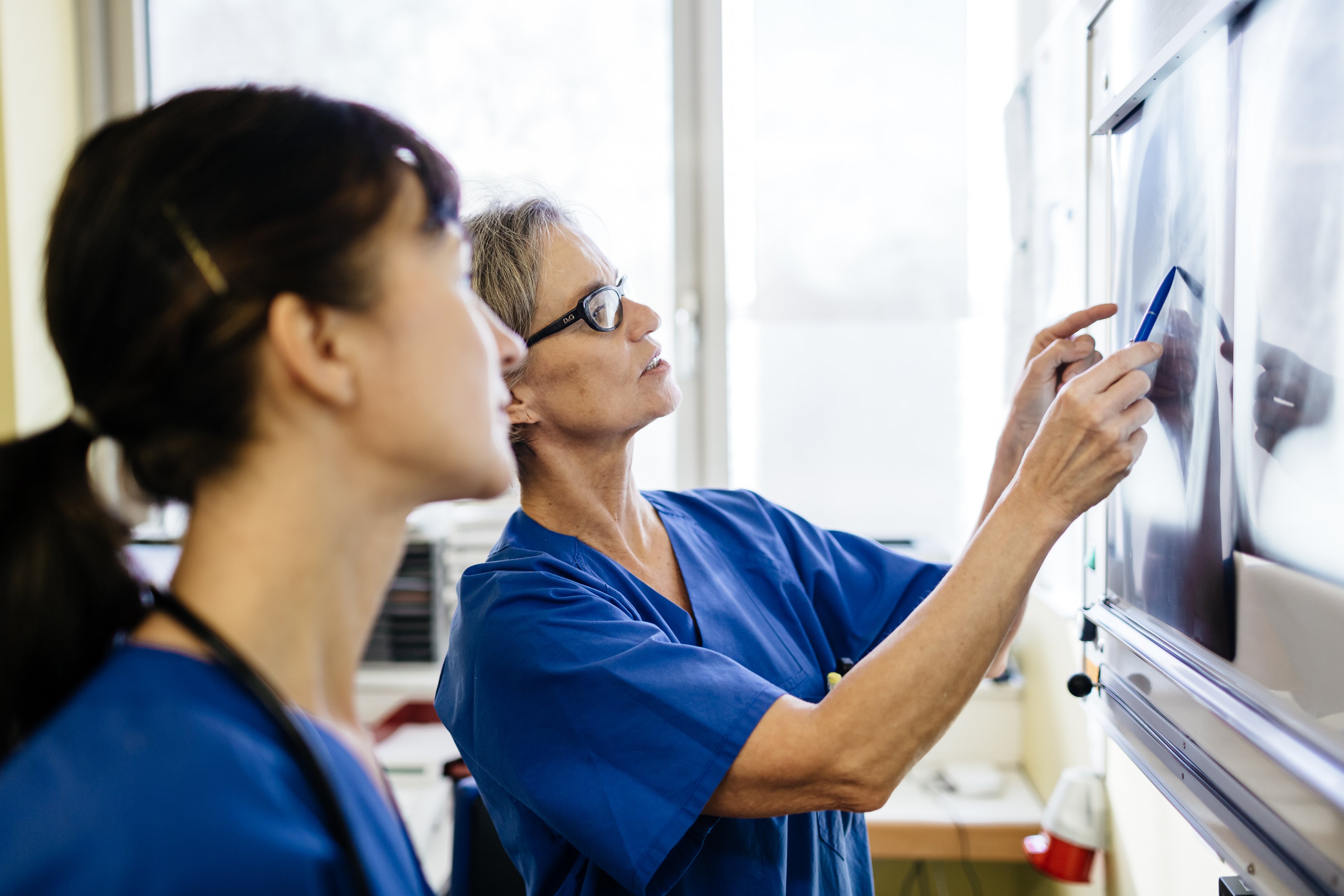 Two female doctors in blue scrubs stand in front of x-ray images, pointing out various elements.