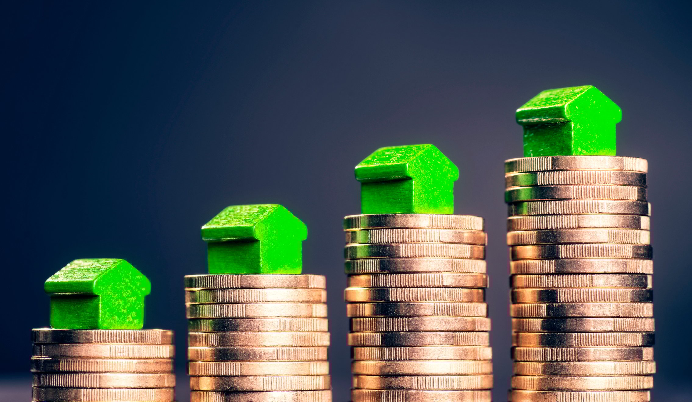 Miniature toy houses are on top of rising stacks of gold coins.