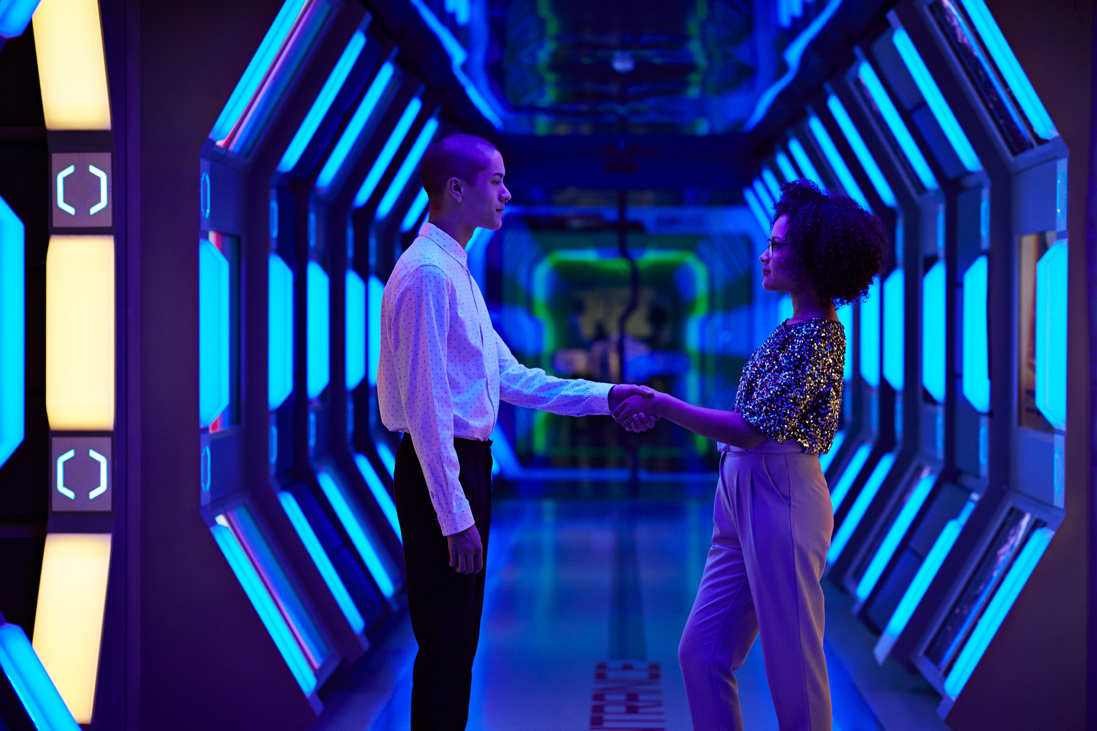 A man and a woman face each other and shake hands in a hallway enveloped by bright blue futuristic lights.