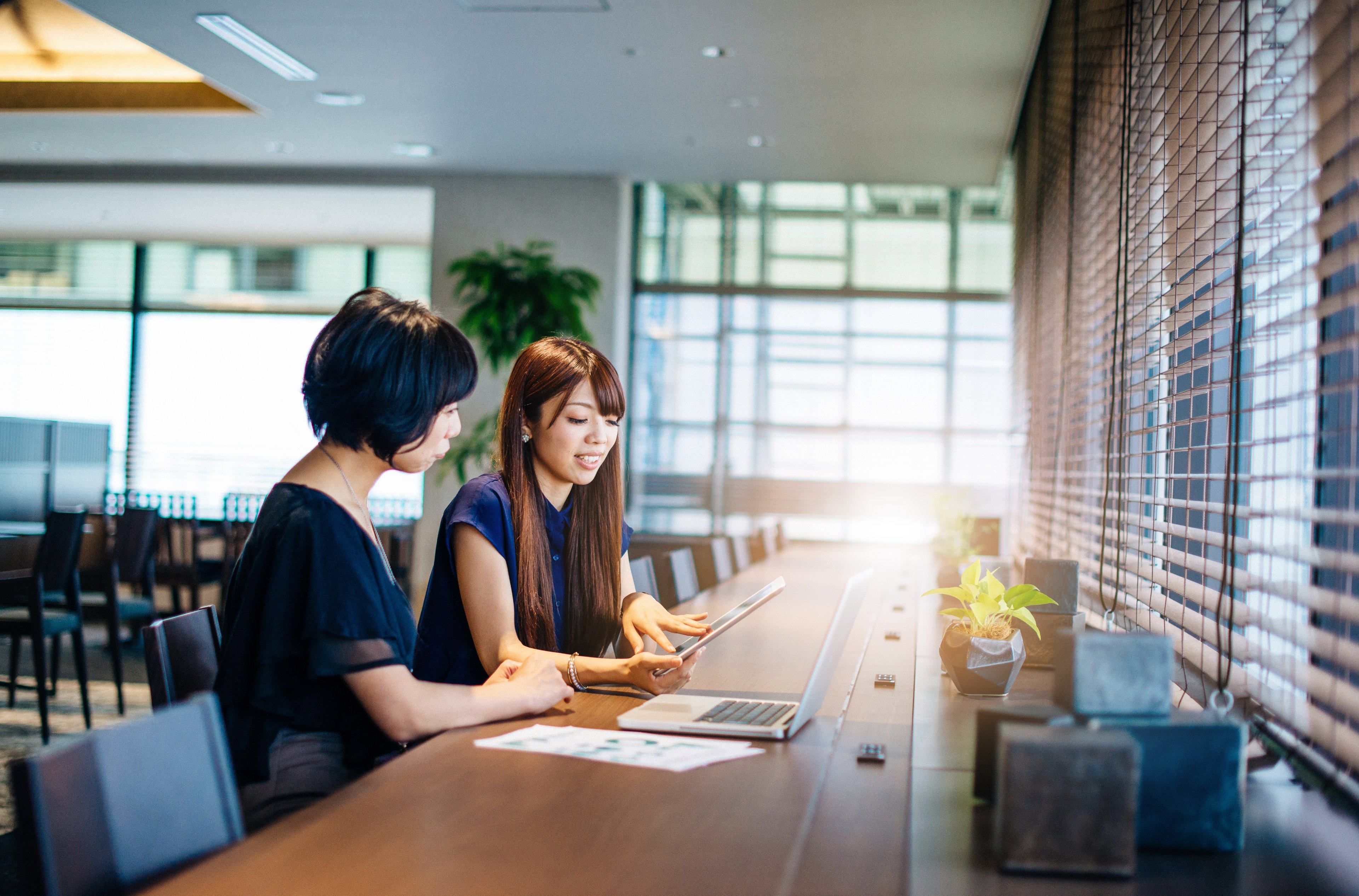 Two women sit at a counter in a large meeting space in front of a laptop computer, with the sunlight shining through the window in the background.