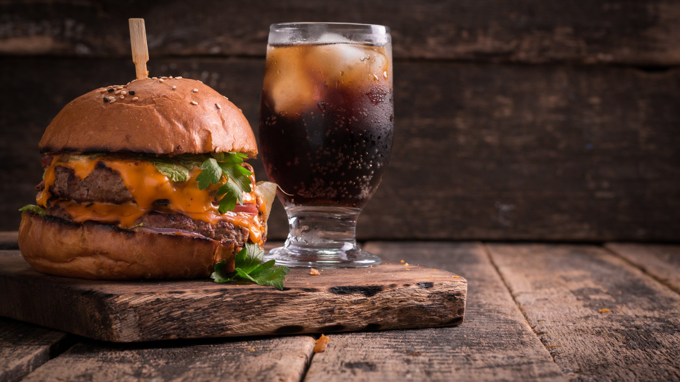 A hamburger next to a Coke in a glass on a wooden slab. 