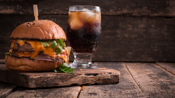 A hamburger next to a Coke in a glass on a wooden slab. 