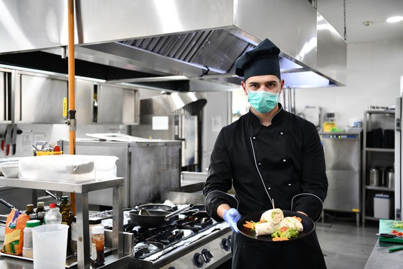 A chef wearing a medical mask holds a plate of food in a commercial kitchen.