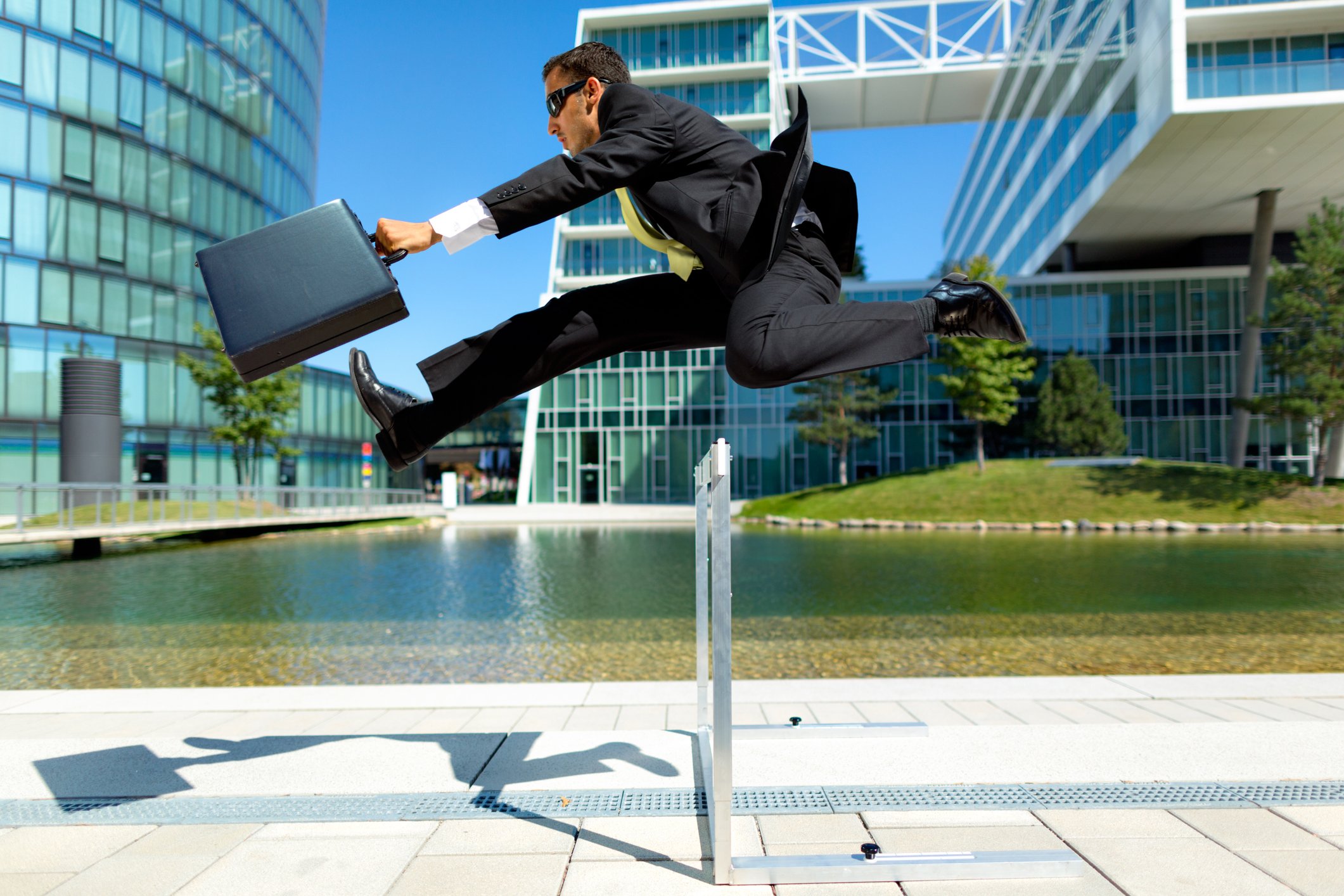 A businessman with a briefcase jumping across a hurdle, with a backdrop of modern buildings.