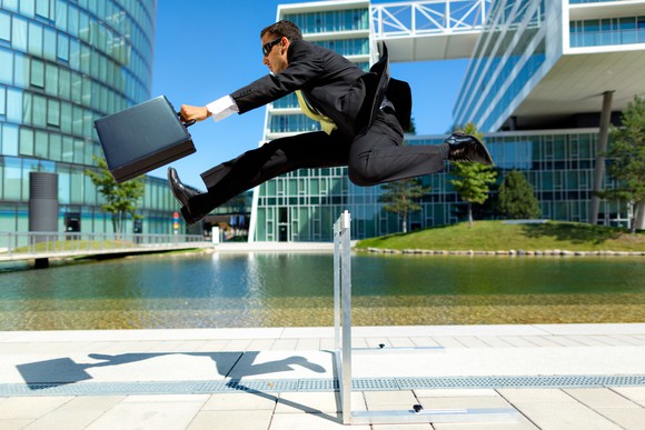 A businessman with a briefcase jumping across a hurdle, with a backdrop of modern buildings.