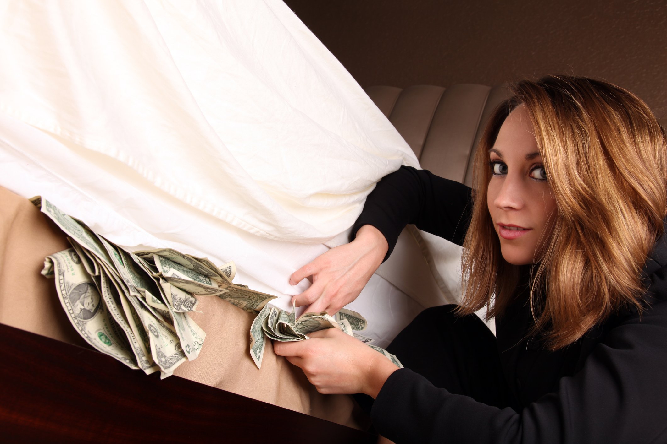 A young woman looks into the camera as she stashes cash under a mattress. 