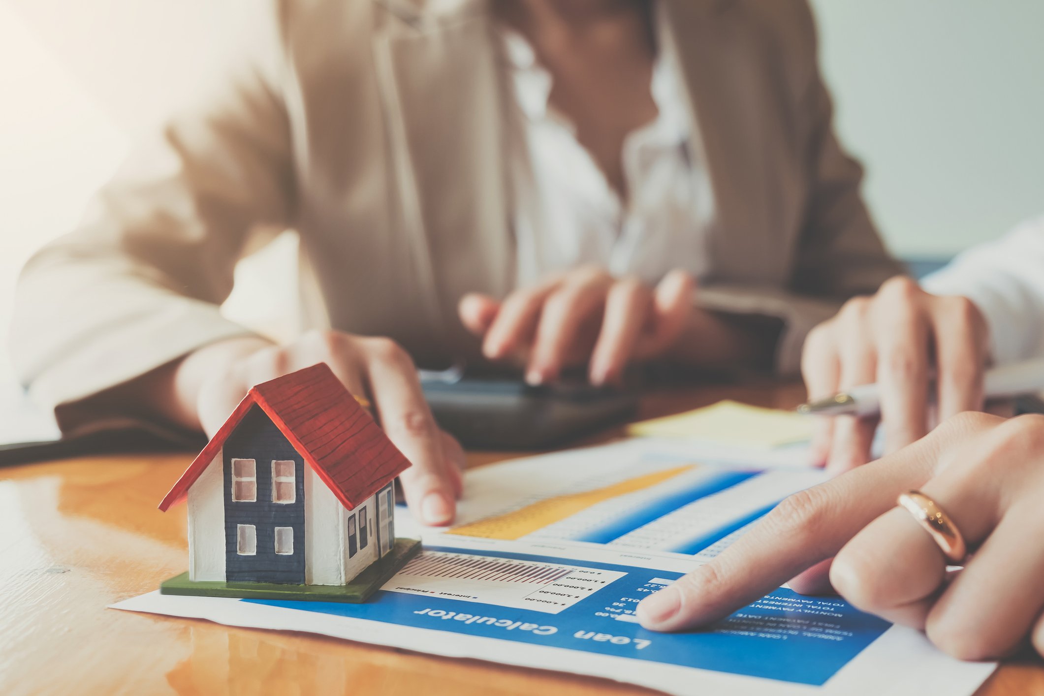 A table with mortgage paperwork and a small house model, as hands point at the paper.