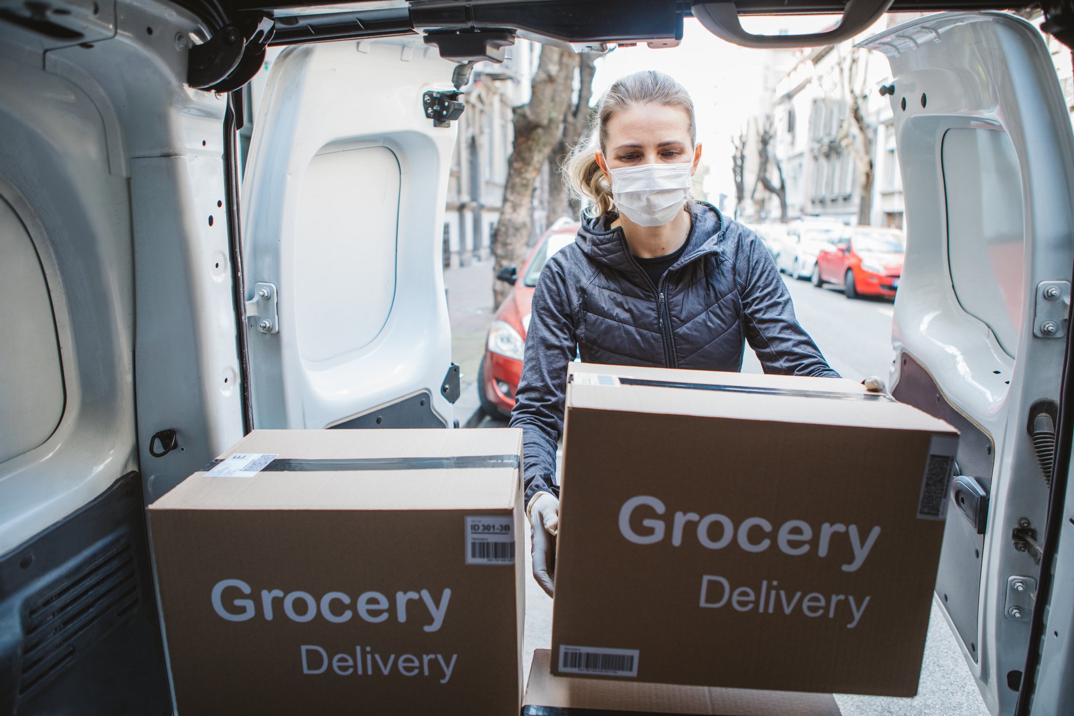 A woman in a mask taking boxes labeled "Grocery Delivery" out of the back of a sprinter van. 