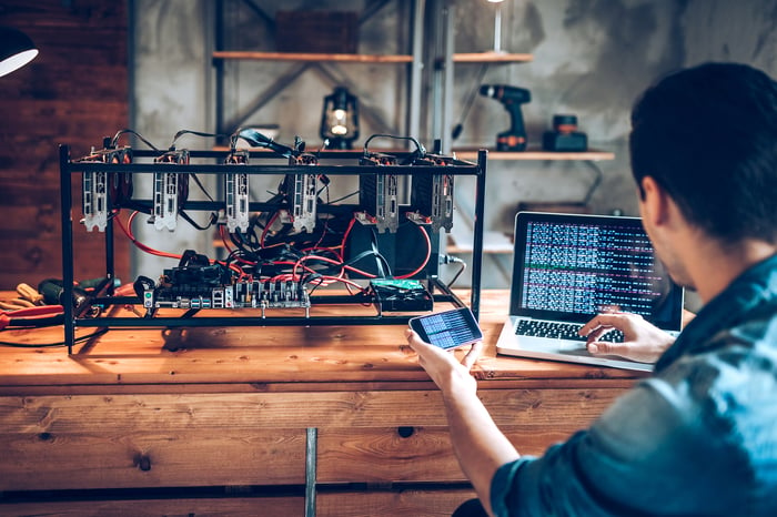 A man at a desk with a laptop operating cryptocurrency mining hardware.