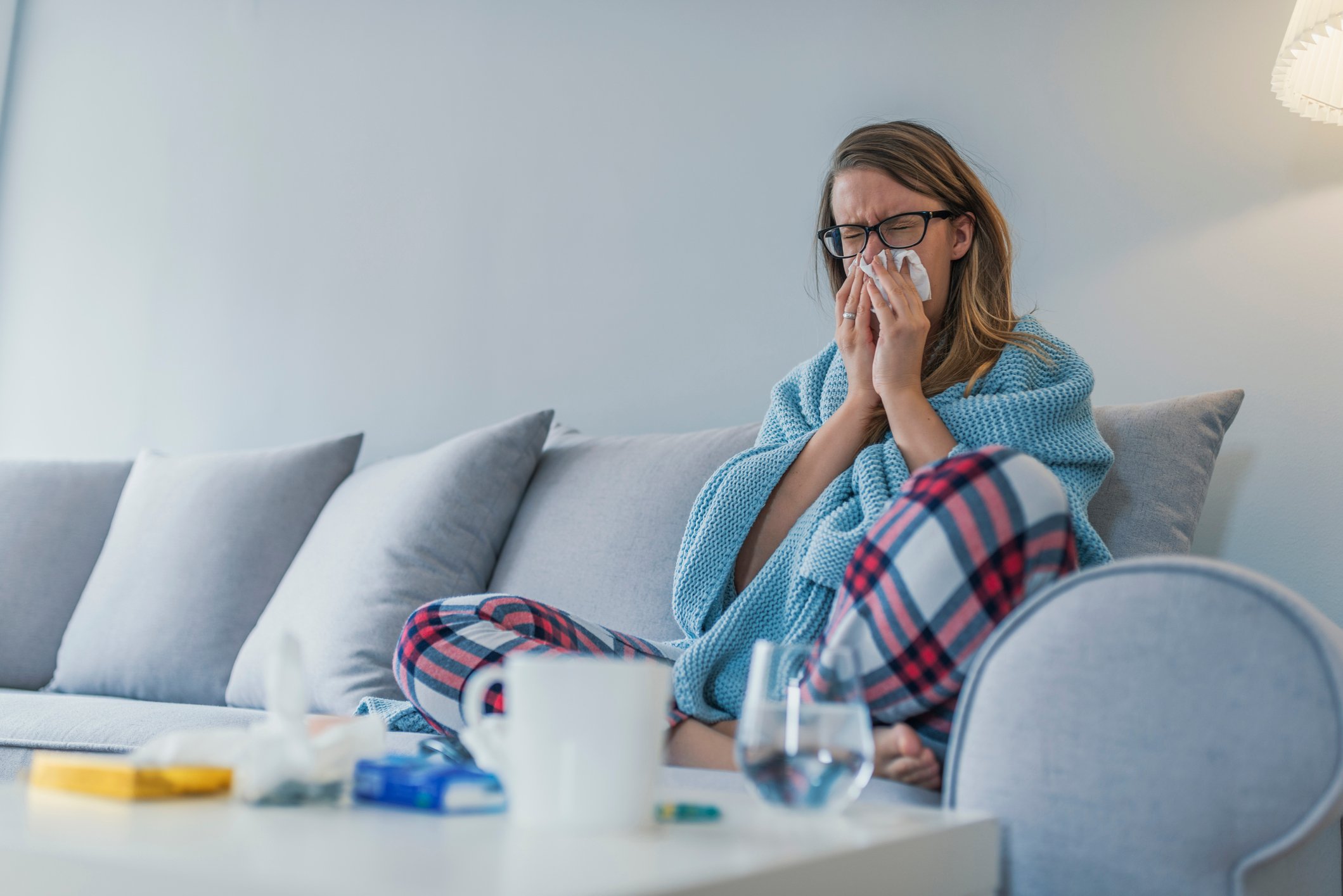 A woman in her pajamas, wrapped in a blanket, sitting on the couch blowing her nose.