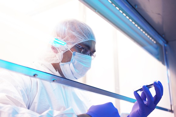 Man wearing a mask, white protective gear, gloves, and glasses investigates materials on a sheet in a laboratory setting.