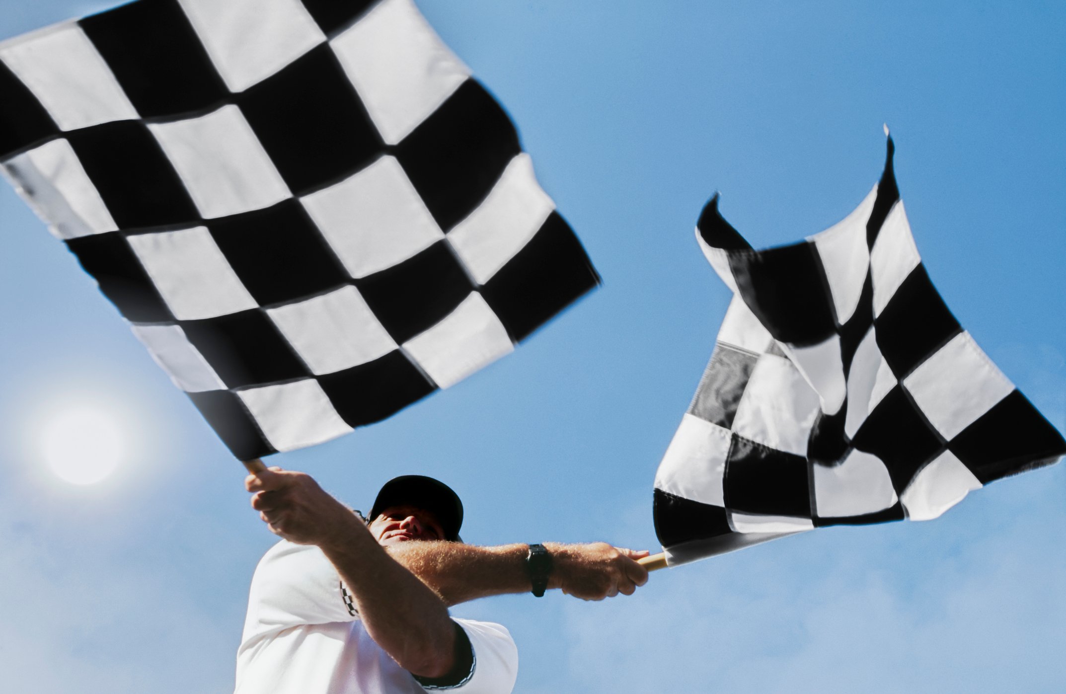 A racetrack official waving two checkered flags against a blue sky.