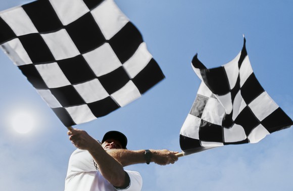 A racetrack official waving two checkered flags against a blue sky.