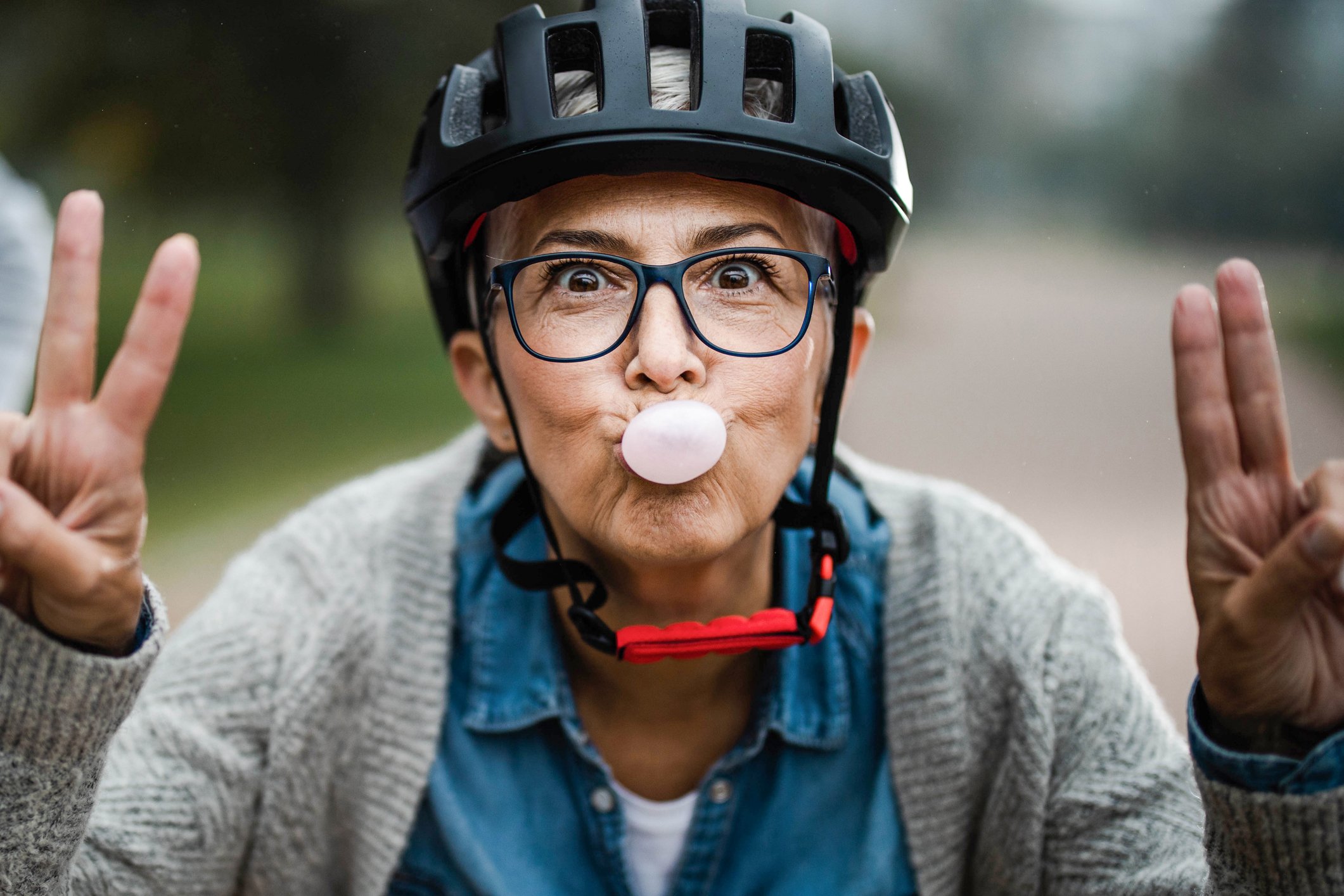 A playful senior woman shows peace signs while blowing bubble gum in a park.
