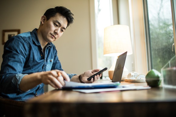Young man holds his cell phone in one hand and flips over a piece of paper with another, sitting at a desk in front of a window in the daylight.
