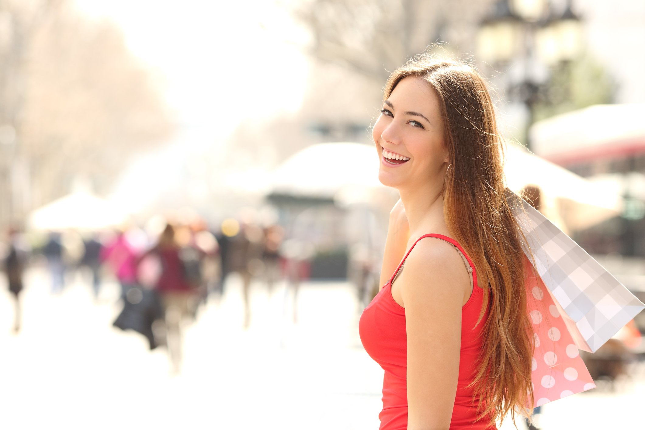 A young woman in a summer dress smiles at the camera, holding several shopping bags over her shoulder.