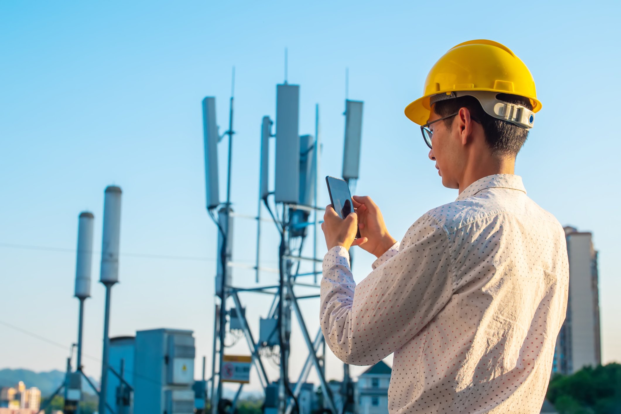 Technician in a hardhat checking his smartphone as he stands near a tower providing service.