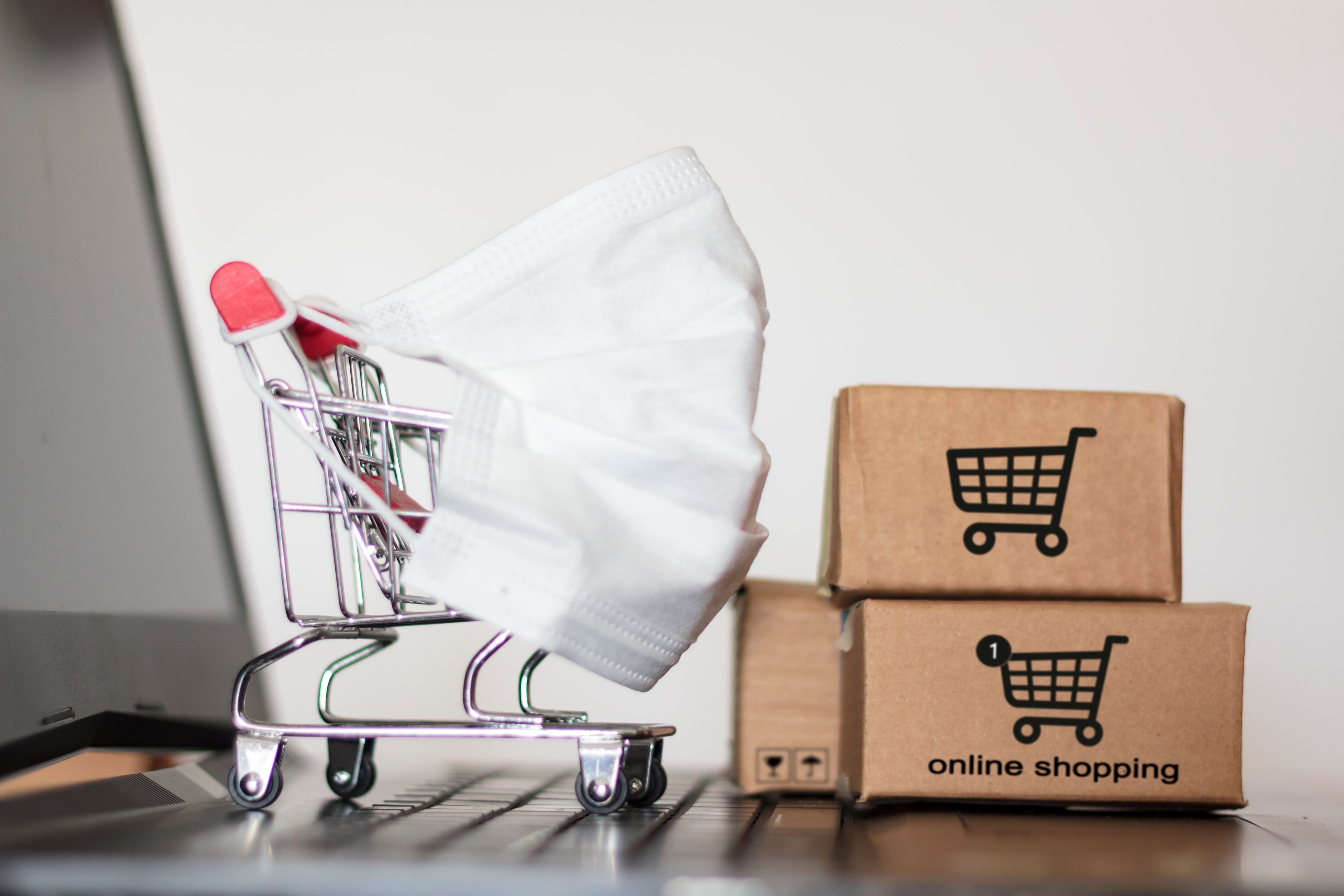 Small grocery cart with face mask on the front of it and shipping boxes sitting on a laptop