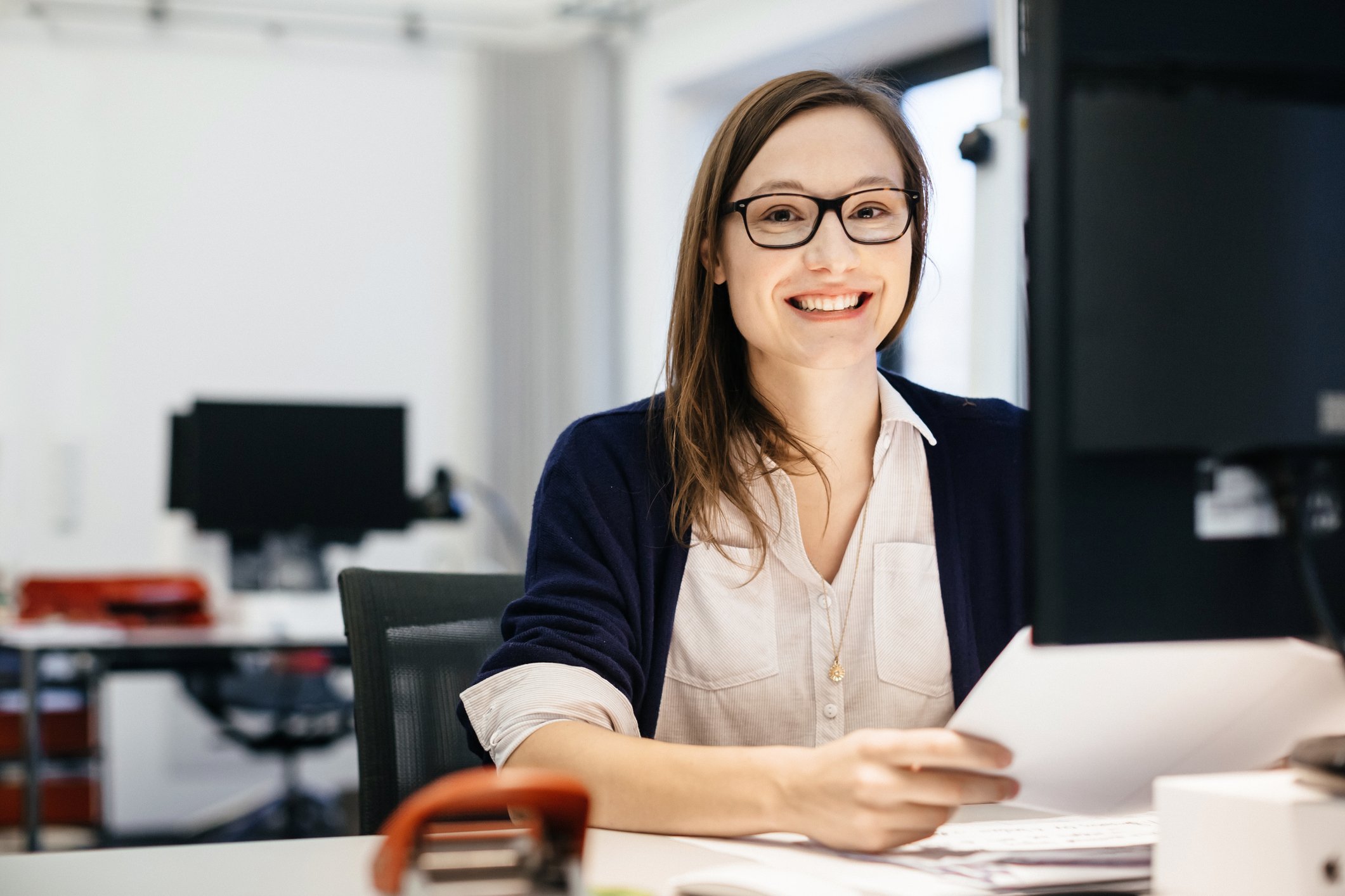 Smiling woman at a desk in front of PC monitor, holding a document.