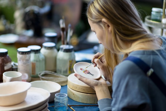 Lady putting finishing touch to a painting on a coaster using a fine paint brush.