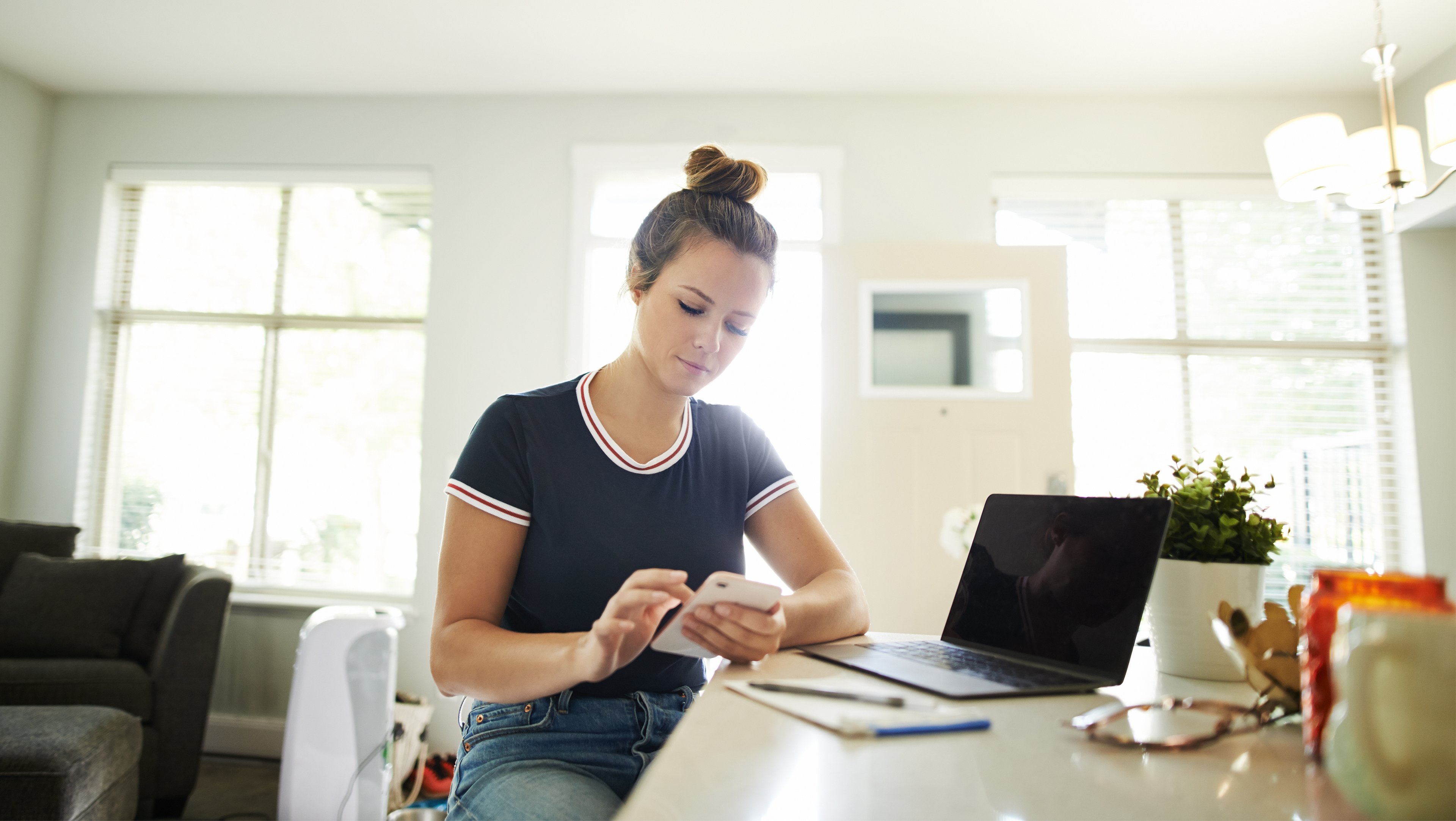 Woman sitting at a countertop looks down at her smartphone, sitting in front of a pad of paper and her laptop computer. 