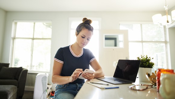 Woman sitting at a countertop looks down at her smartphone, sitting in front of a pad of paper and her laptop computer. 