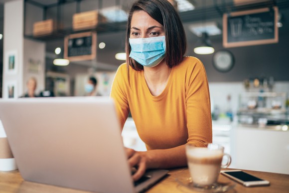 A woman in a medical mask works on her laptop in a coffee shop.