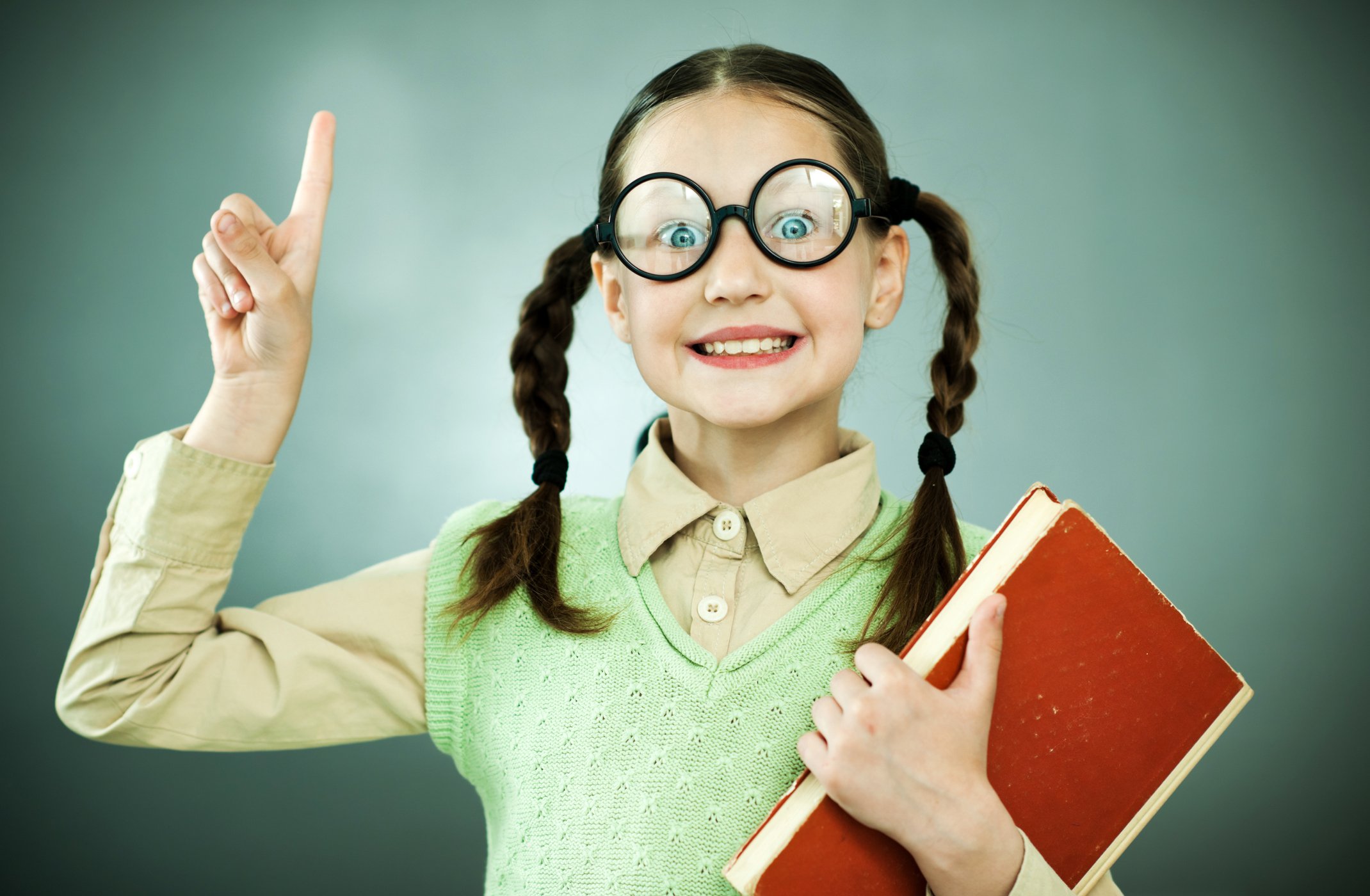 A young girl in glasses is holding up one finger and carrying a book.