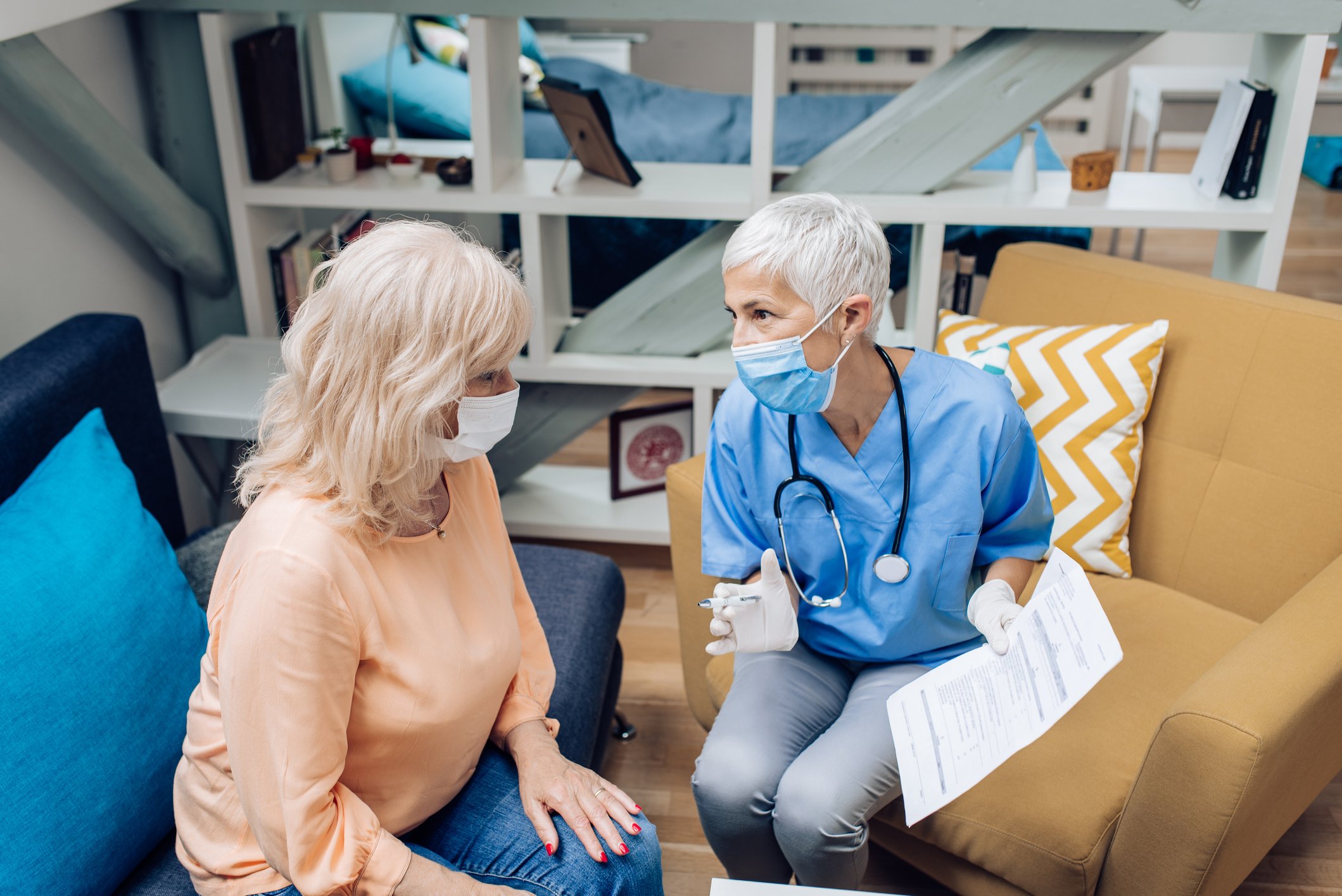 One woman, a patient wearing a mask, sits on a blue couch opposite a woman healthcare provider, also masked and gloved, who shows her medical forms. 
