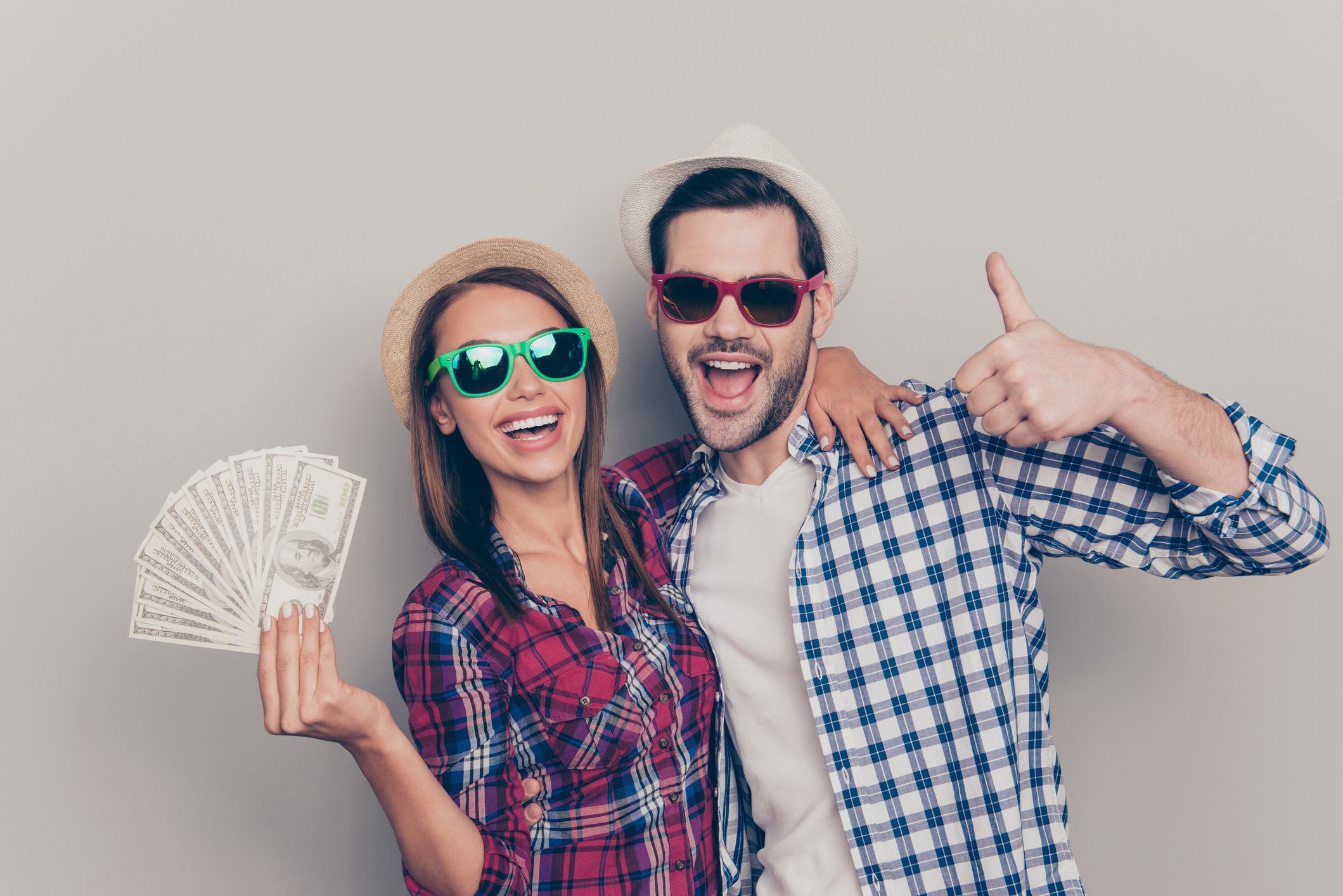 A young man giving a thumbs up sign stands beside a young woman holding a fan of cash. 