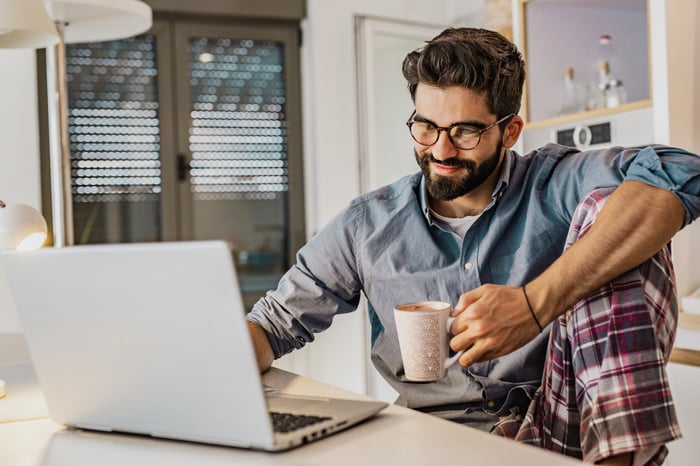 A young man wearing pajama pants works from home at his kitchen counter.