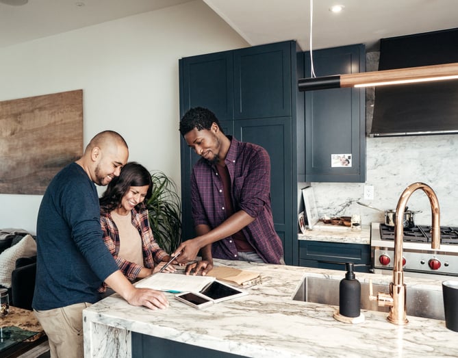 Young man appears to assist a young couple, as they stand by a kitchen counter in front of notebooks, a smartphone, and a tablet