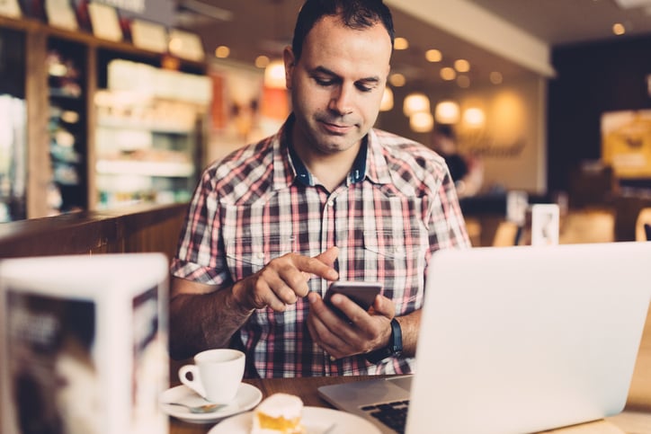 Man types on smartphone while sitting at a cafe table in front of his laptop computer and a cup of coffee.