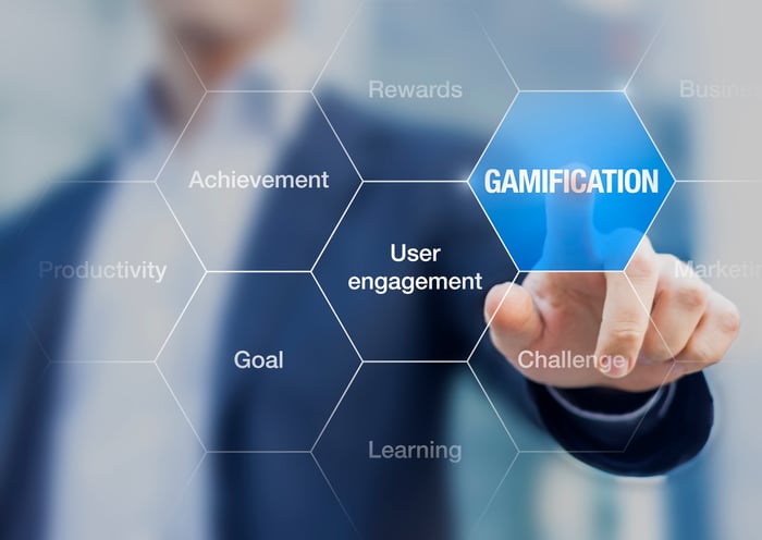 A businessman in a dark blue suit pointing to a medium blue electronic hexagon labeled "gamification." 