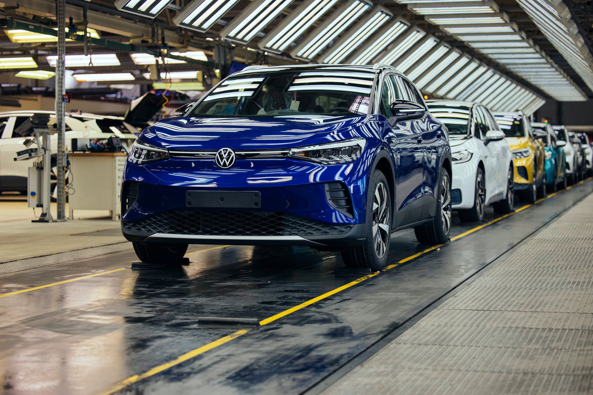 A blue Volkswagen ID.4, an electric SUV, leads a line of VW cars at the end of a factory assembly line. 