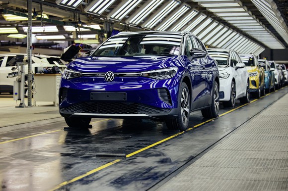 A blue Volkswagen ID.4, an electric SUV, leads a line of VW cars at the end of a factory assembly line. 