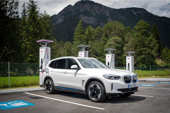 White BMW vehicle at charging station with conifers and mountains in the background.