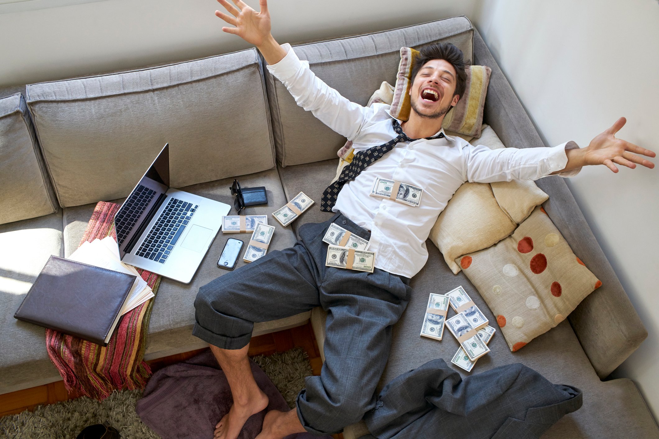 man sitting back on couch celebrating with piles of cash