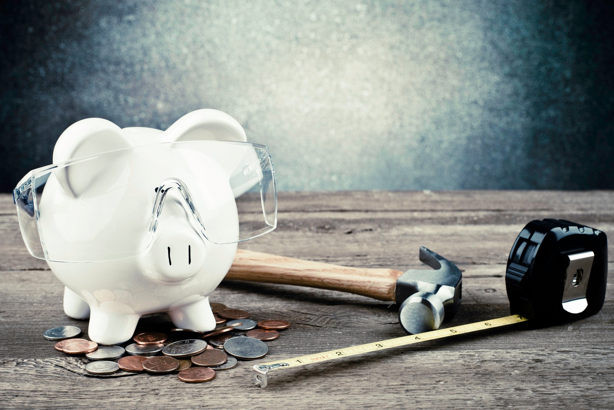 A piggy bank with safety goggles on surrounded by a hammer, a tape measure, and loose change. 