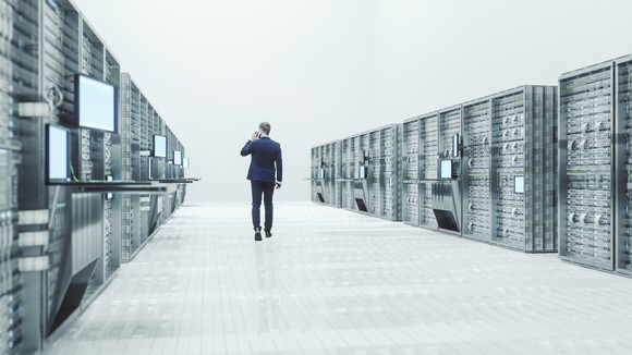 Man walsk between two rows of server racks in a big open white room. 